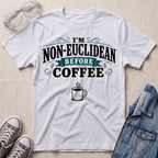 A white graphic t-shirt laid flat on a light beige textured background, styled with safety glasses, gray sneakers, and a denim jacket. The t-shirt features a graphic that reads 'I'M NON-EUCLIDEAN BEFORE COFFEE' in black and teal lettering, with a small coffee cup illustration below.