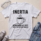 A white graphic t-shirt laid flat on a light beige fabric background, styled with safety goggles, gray sneakers, and a blue denim jacket. The t-shirt features a black graphic of a steaming coffee mug with the text 'INERTIA REMAINING AT REST UNTIL THE CAFFEINE KICKS IN'.