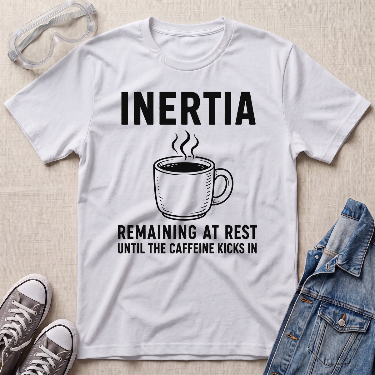 A white graphic t-shirt laid flat on a light beige fabric background, styled with safety goggles, gray sneakers, and a blue denim jacket. The t-shirt features a black graphic of a steaming coffee mug with the text 'INERTIA REMAINING AT REST UNTIL THE CAFFEINE KICKS IN'.
