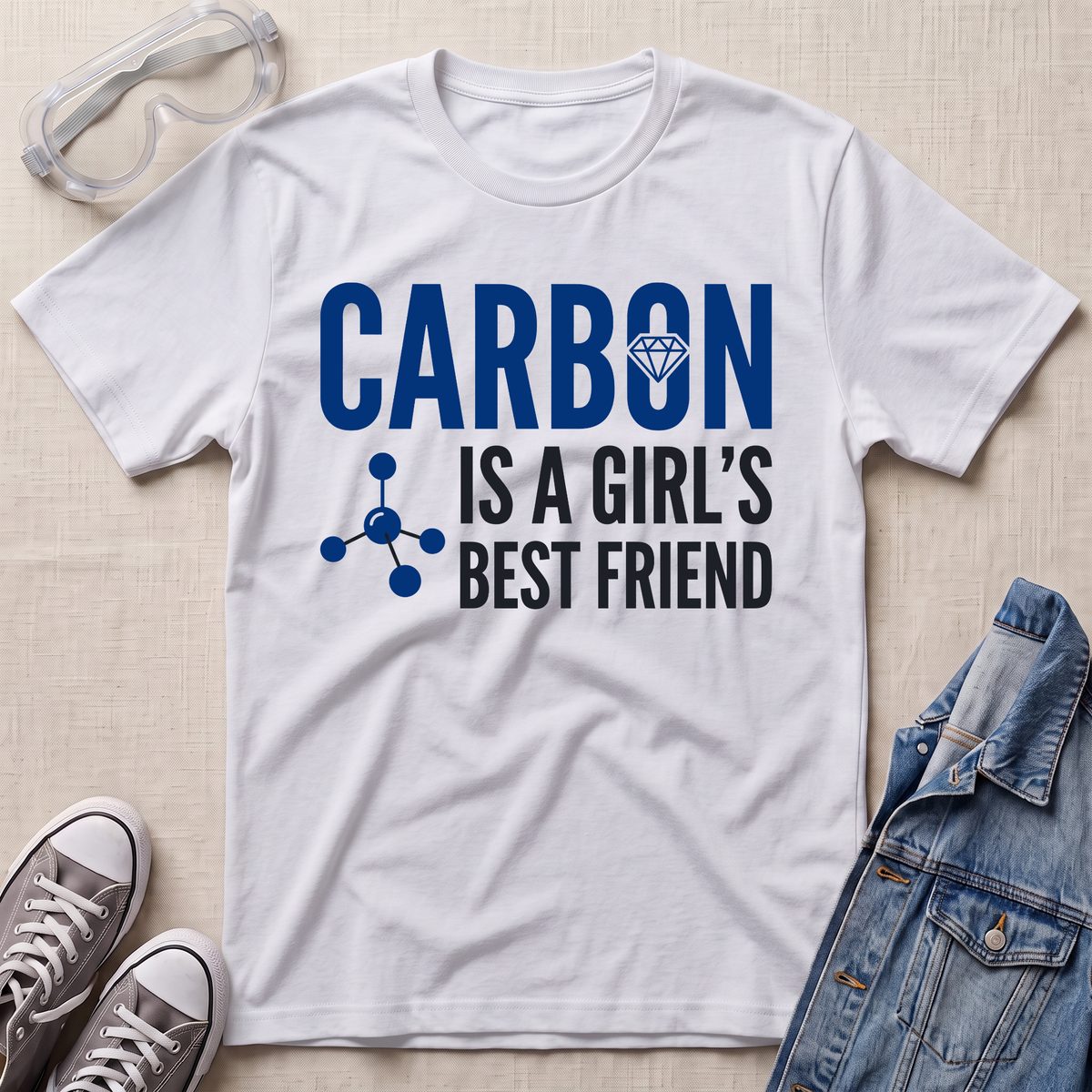 A white short-sleeve graphic T-shirt laid flat on a light fabric surface, styled with safety goggles, gray sneakers, and a denim jacket. The shirt has large blue text reading 'CARBON' with a small diamond icon, and black text reading 'IS A GIRL'S BEST FRIEND' with a blue molecule graphic.