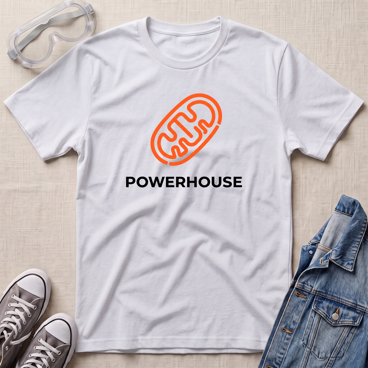 A white short-sleeve t-shirt laid flat on a light beige textured surface, featuring an orange mitochondrial outline graphic above the word 'POWERHOUSE' in black. Styled flat-lay props include clear safety goggles, gray sneakers, and a blue denim jacket.