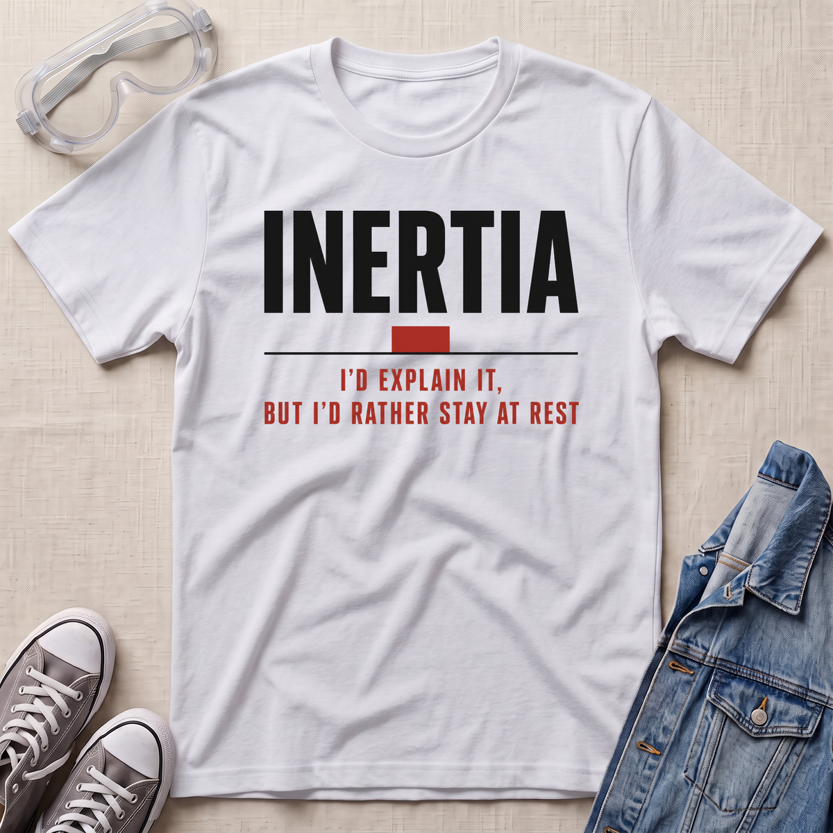 A white graphic t-shirt laid flat on a light beige textured background, styled with safety goggles, gray sneakers, and a denim jacket. The t-shirt features the word 'INERTIA' in bold black letters, with a red block and red text underneath reading 'I'D EXPLAIN IT, BUT I'D RATHER STAY AT REST'.