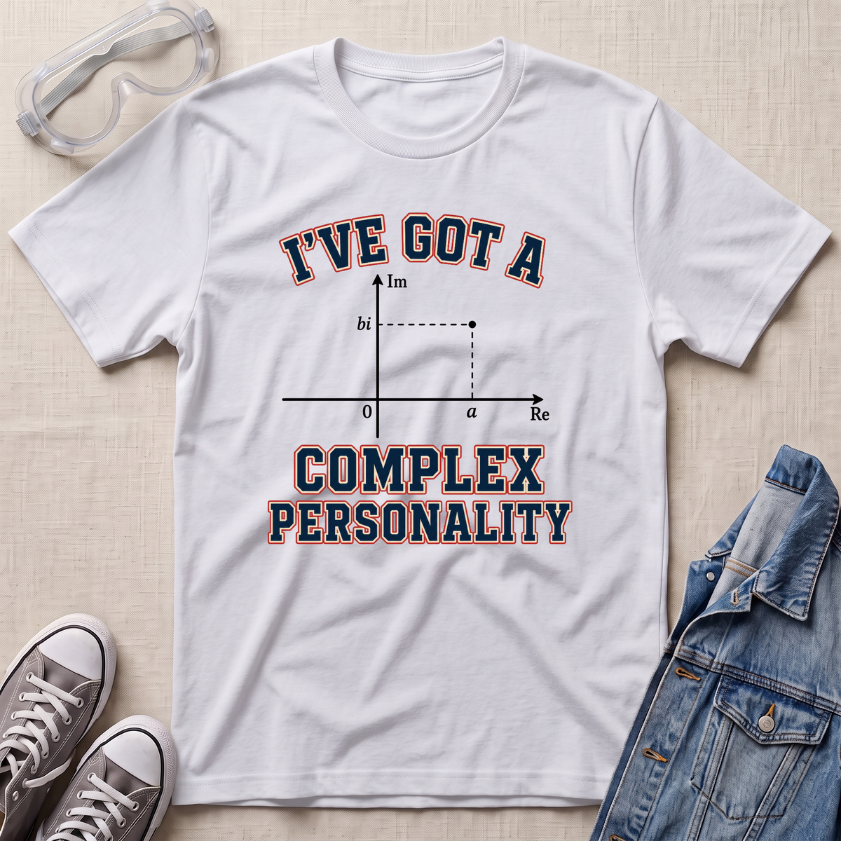 A white graphic t-shirt laid flat on a beige textured surface, styled with safety goggles, gray sneakers, and a denim jacket. The t-shirt features a graphic of a complex plane with the text 'I'VE GOT A COMPLEX PERSONALITY' in navy blue with a red outline.