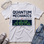 A white graphic t-shirt laid flat on a light beige textured background, styled with clear safety goggles, gray sneakers, and a blue denim jacket. The t-shirt features a dark blue graphic that reads 'QUANTUM MECHANICS' above a green and blue retro computer screen saying 'IF YOU THINK YOU UNDERSTAND IT, YOU'RE DOING IT WRONG.
