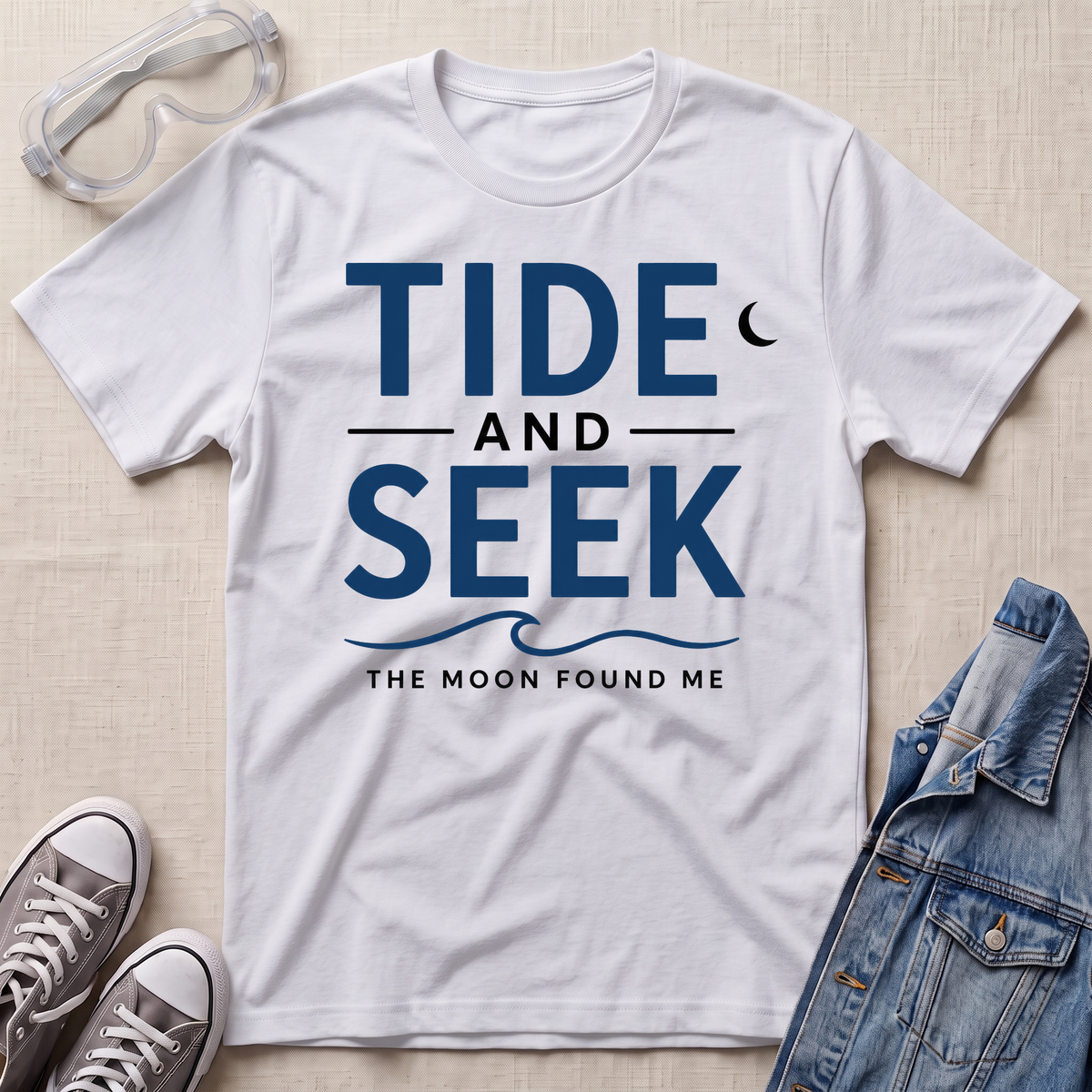 A white short-sleeve graphic T-shirt laid flat on a light beige textured surface, with safety glasses, gray sneakers, and a folded denim jacket around it. The shirt has large navy text reading 'TIDE AND SEEK' with smaller text 'THE MOON FOUND ME' and a crescent moon graphic.