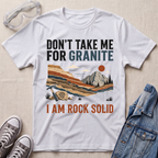 A white graphic t-shirt laid flat on a textured beige surface, surrounded by styled props including clear safety glasses, gray sneakers, and a blue denim jacket. The t-shirt features a colorful mountain landscape illustration with the text 'DON'T TAKE ME FOR GRANITE' above and 'I AM ROCK SOLID' below.