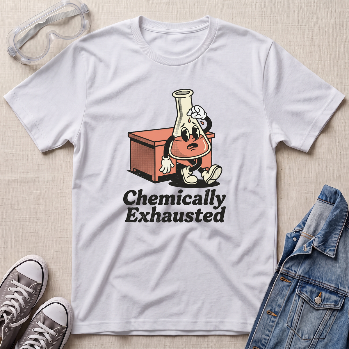 A white short-sleeve graphic T-shirt laid flat on a light fabric background, styled with safety goggles, gray sneakers, and a denim jacket. The shirt features a cartoon lab flask character with a flower on its head sitting on an orange cabinet, above the text 'Chemically Exhausted'.
