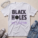 A white graphic t-shirt laid flat on a light beige textured background, styled with clear safety goggles, gray sneakers, and a blue denim jacket. The t-shirt features a bold graphic that reads 'BLACK HOLES SUCK AT EVERYTHING' in black and purple text.