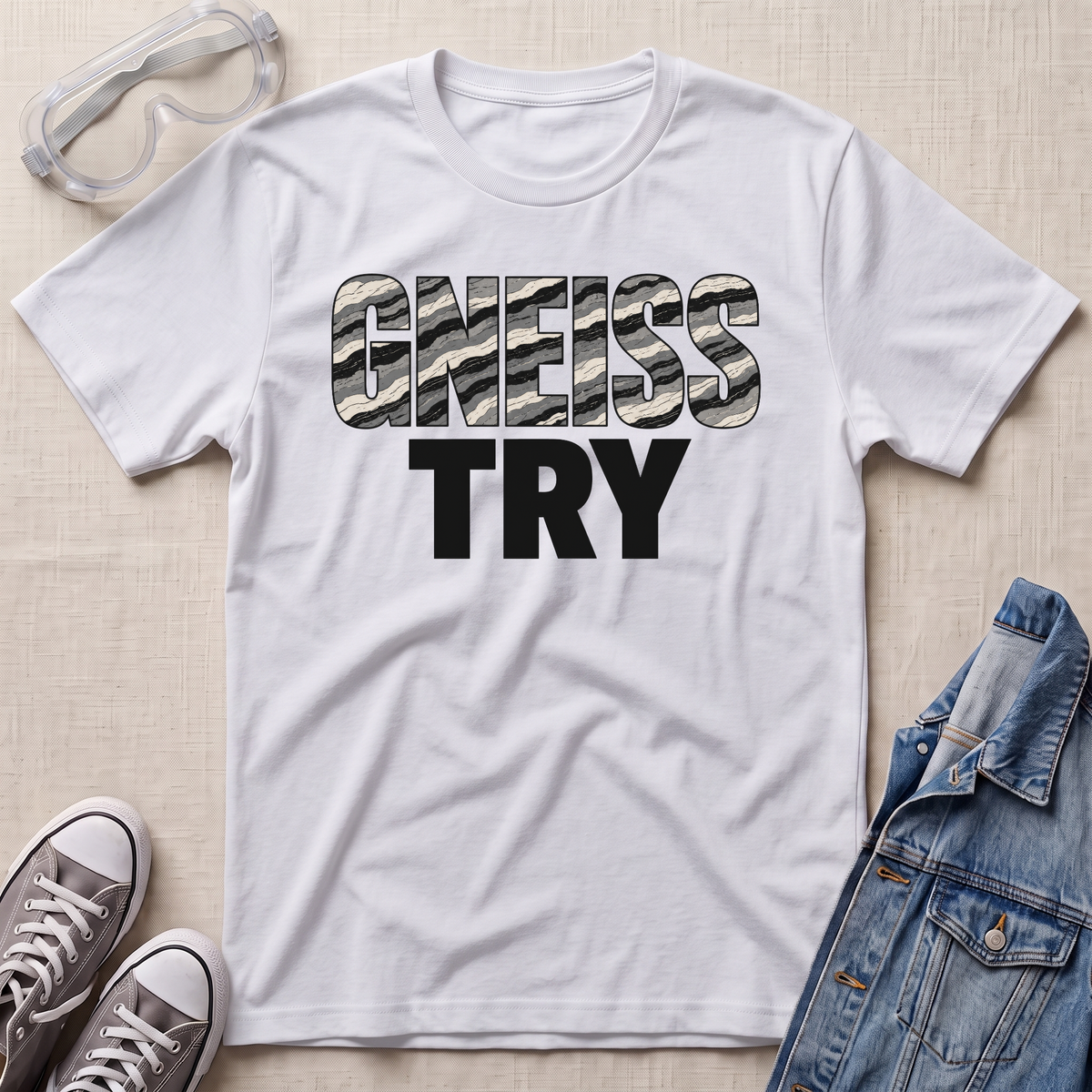 A white short-sleeve graphic T-shirt laid flat on a light beige textured surface, styled with clear safety goggles, gray sneakers, and a blue denim jacket. The shirt has large text reading 'GNEISS TRY' across the chest, with 'GNEISS' filled with a black-and-white zebra pattern and 'TRY' in solid black.