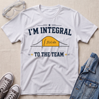 A white graphic t-shirt laid flat on a light beige textured surface, surrounded by styling props including clear safety glasses, gray sneakers, and a blue denim jacket. The t-shirt features a dark navy blue graphic that reads 'I'M INTEGRAL TO THE TEAM' with a yellow integral curve diagram in the center.