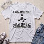 A white graphic t-shirt featuring a black printed text design that reads 'I AM A MOLECULE ASK ME ABOUT MY CONFORMATION' with a simple molecule diagram. The t-shirt is displayed in a flat-lay arrangement on a light beige textured background, surrounded by props including safety goggles, gray sneakers, and a blue denim jacket.