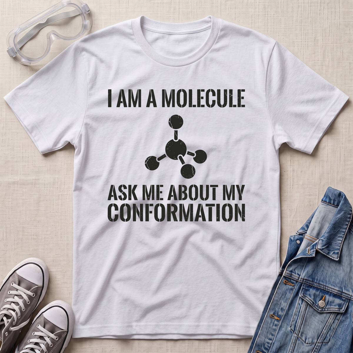 A white graphic t-shirt featuring a black printed text design that reads 'I AM A MOLECULE ASK ME ABOUT MY CONFORMATION' with a simple molecule diagram. The t-shirt is displayed in a flat-lay arrangement on a light beige textured background, surrounded by props including safety goggles, gray sneakers, and a blue denim jacket.