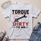 A white graphic T-shirt laid flat on a light beige fabric background, styled with safety glasses, gray sneakers, and a blue denim jacket. The shirt features a black and red graphic that reads 'TORQUE DIRTY TO ME' with a wrench and torque arrow design.