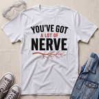 A white graphic t-shirt laid flat on a light beige textured surface, surrounded by styling props including clear safety goggles, gray sneakers, and a blue denim jacket. The t-shirt features the text 'YOU'VE GOT A LOT OF NERVE' in black and red lettering with a red branch graphic underneath.
